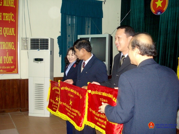 Đồng chí Nguyễn Văn A (người đứng giữa) nhận cờ thi đua của Viện KSND tối cao tặng tập thể dẫn đầu phong trào thi đua khối năm 2008. (một năm trước thời điểm nghỉ hưu)